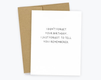 May include: A white greeting card with the text "I DIDN'T FORGET YOUR BIRTHDAY. I JUST FORGOT TO TELL YOU I REMEMBERED." is placed in front of a brown envelope. The card and envelope are on a white background.