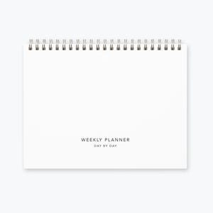 May include: A white weekly planner with a spiral binding. The planner has the text "WEEKLY PLANNER DAY BY DAY" printed on the front.