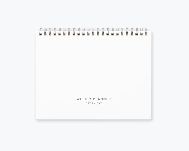 May include: A white weekly planner with a spiral binding. The planner has the text "WEEKLY PLANNER DAY BY DAY" printed on the front.