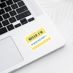 May include: A yellow sticker with the text "HELLO I'M" in teal and "trying my best" in teal, with a small heart. The sticker is on a white laptop.