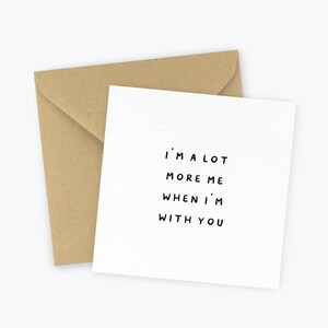 May include: A white greeting card with the text "I'M A LOT MORE ME WHEN I'M WITH YOU" in black font, accompanied by a brown envelope. The card and envelope are set against a white background.