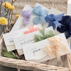 May include: A set of six white envelopes with different colored ribbons tied in bows. The envelopes have text that reads "A note to my..." followed by "mom", "maid of honor", and "groom".