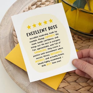 May include: A white card with the text "EXCELLENT BOSS" and a yellow envelope. The card has a yellow oval with text that describes the boss's qualities. The card is held by a person's hand.