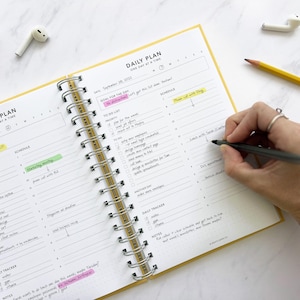 May include: An open daily planner with a yellow cover and a spiral binding. The planner has sections for goals, to-do lists, and schedules. The pages are filled with handwritten notes and checkmarks. A hand is writing in the planner with a black pen.