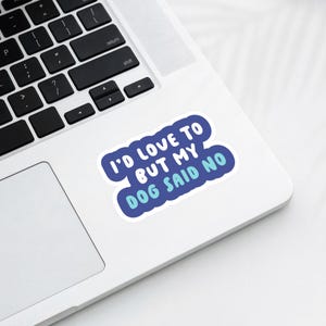 May include: A blue and white sticker with the text "I'D LOVE TO BUT MY DOG SAID NO" in a playful font. The sticker is on a white laptop, with the keyboard visible in the background. The sticker has a blue border.