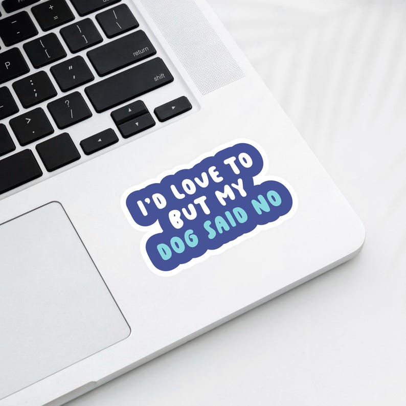 May include: A blue and white sticker with the text "I'D LOVE TO BUT MY DOG SAID NO" in a playful font. The sticker is on a white laptop, with the keyboard visible in the background. The sticker has a blue border.