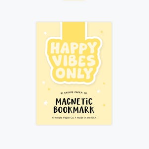 May include: A yellow magnetic bookmark with the text "HAPPY VIBES ONLY" in a bubble font. The words "MAGNETIC BOOKMARK" are below the text. The bookmark has a light yellow background with small star accents.