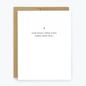 May include: A white greeting card with the text "SOME ANGELS HAVE WINGS, OTHERS HAVE TAILS." in black, handwritten-style font. A small black heart is above the text. The card is inside a brown envelope.