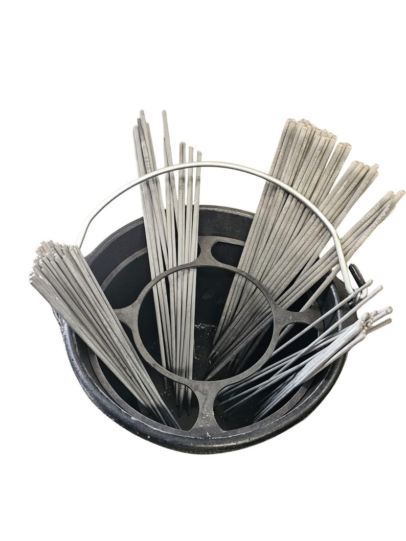 May include: A black metal bucket filled with numerous gray welding rods. The rods are arranged in bundles and held in place by a metal frame. The image is taken from a high angle, showing the contents of the bucket.