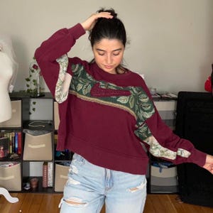 May include: Burgundy-colored sweatshirt with a floral design on the front and sleeves. The Nike logo is embroidered in a darker red. The sweatshirt is paired with light blue jeans.
