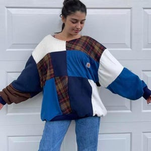 May include: A patchwork sweater featuring a mix of fabrics and colors. The design includes navy blue, white, brown, and plaid panels. The sweater has a relaxed fit and long sleeves. A small logo is visible on the front.