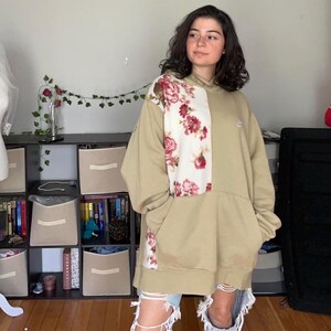 May include: Oversized tan hoodie with a floral panel on the front and sleeve. The floral panel features a white background with pink and red roses. The hoodie has a kangaroo pocket and a small Nike logo.