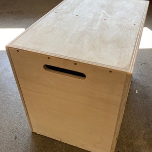 May include: A wooden box with a handle on the front. The box is made of light-colored wood and has a smooth finish.