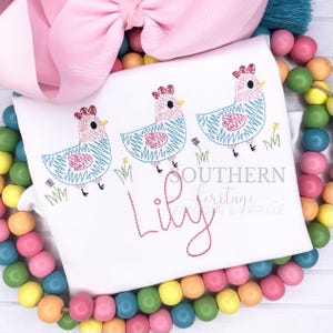 May include: White t-shirt featuring three embroidered chickens with blue and pink details, walking on grass. The name "Lily" is embroidered in pink script. A large pink bow and colorful beads are in the background.