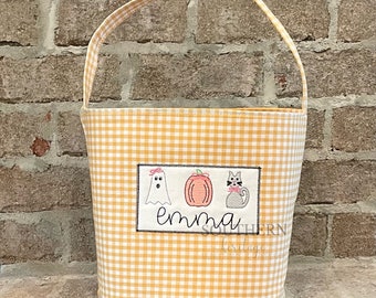Pode incluir: Um balde de Halloween xadrez amarelo e branco com uma alça. O balde tem um patch retangular com imagens bordadas de um fantasma, abóbora e gato, com o nome "Emma" abaixo. O balde é projetado para doces ou travessuras.