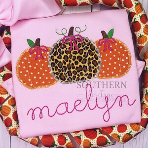 May include: Pink t-shirt with three embroidered pumpkins. Two pumpkins are orange with white polka dots, and one is leopard print. The name "maelyn" is embroidered in pink script below the pumpkins. The text "SOUTHERN Heritage" is also visible.