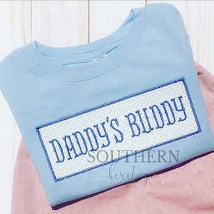 May include: A light blue t-shirt with the embroidered text "DADDY'S BUDDY" in blue, framed by a white rectangle with a textured pattern. The shirt is folded on a pink surface. The text "SOUTHERN heritage MONOGRAMS & APPLIQUE" is below the main text.