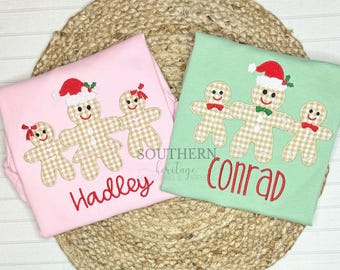 May include: Two long-sleeved shirts, one pink and one green, with gingerbread family designs. The gingerbread people have gingham bodies and wear Santa hats. The pink shirt says "Hadley" in red script, and the green shirt says "Conrad".