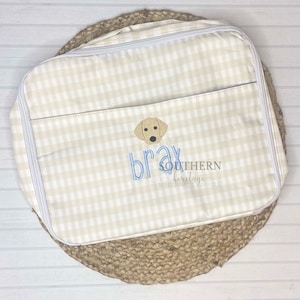 May include: Cream and white gingham travel bag with a light brown dog face applique and the name "Bray" embroidered in blue. The bag has a zippered closure and a front pocket. The text "SOUTHERN heritage MONOGRAMS & APPLIQUE" is also embroidered.