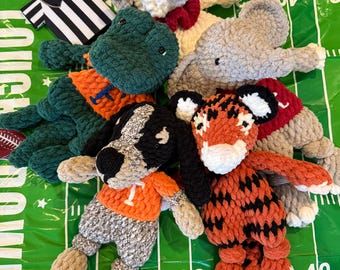 May include: A collection of crocheted stuffed animals in the shape of a tiger, elephant, alligator, dog, and a white animal wearing a red bow tie. The stuffed animals are on a green and white football field.