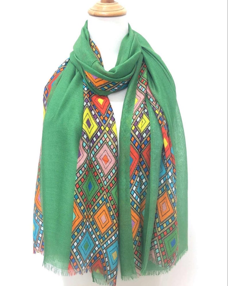 May include: A green scarf with a colourful geometric diamond pattern. The scarf is draped over a mannequin, showcasing the vibrant design. The edges of the scarf have a fringed detail. The diamond pattern includes shades of orange, yellow, pink, and blue.