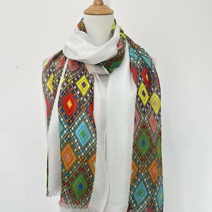 May include: A white scarf with a colorful geometric pattern. The pattern features diamonds in red, yellow, blue, and green. The scarf is displayed on a mannequin.