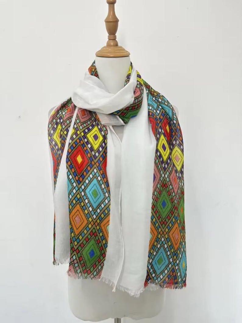 May include: A white scarf with a colorful geometric pattern. The pattern features diamonds in red, yellow, blue, and green. The scarf is displayed on a mannequin.