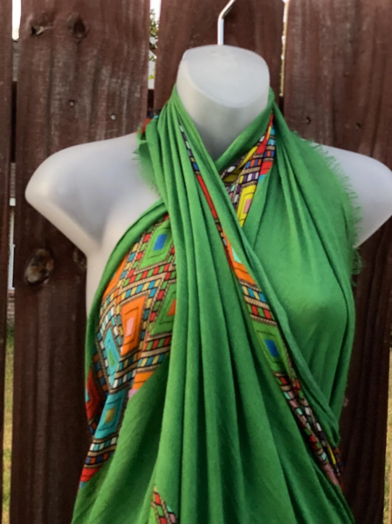 May include: A bright green scarf with a geometric pattern in orange, blue, yellow, and red. The scarf is draped over a mannequin. The design is visible on the edges of the scarf, adding a splash of colour to the green fabric.