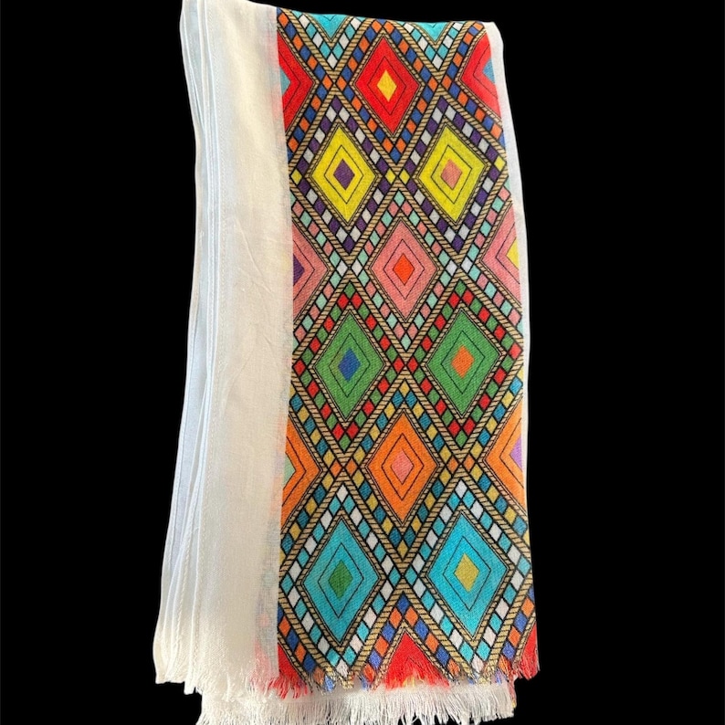 May include: A white scarf with a colorful geometric diamond pattern. The pattern includes red, yellow, pink, green, orange, and blue diamonds. The scarf has a fringed edge with red accents.