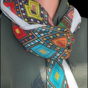 May include: A colorful scarf with a geometric pattern in red, orange, yellow, blue, and purple is knotted around a neck. The scarf has a white border and is draped over a green hooded sweatshirt. The pattern includes diamond and square shapes.