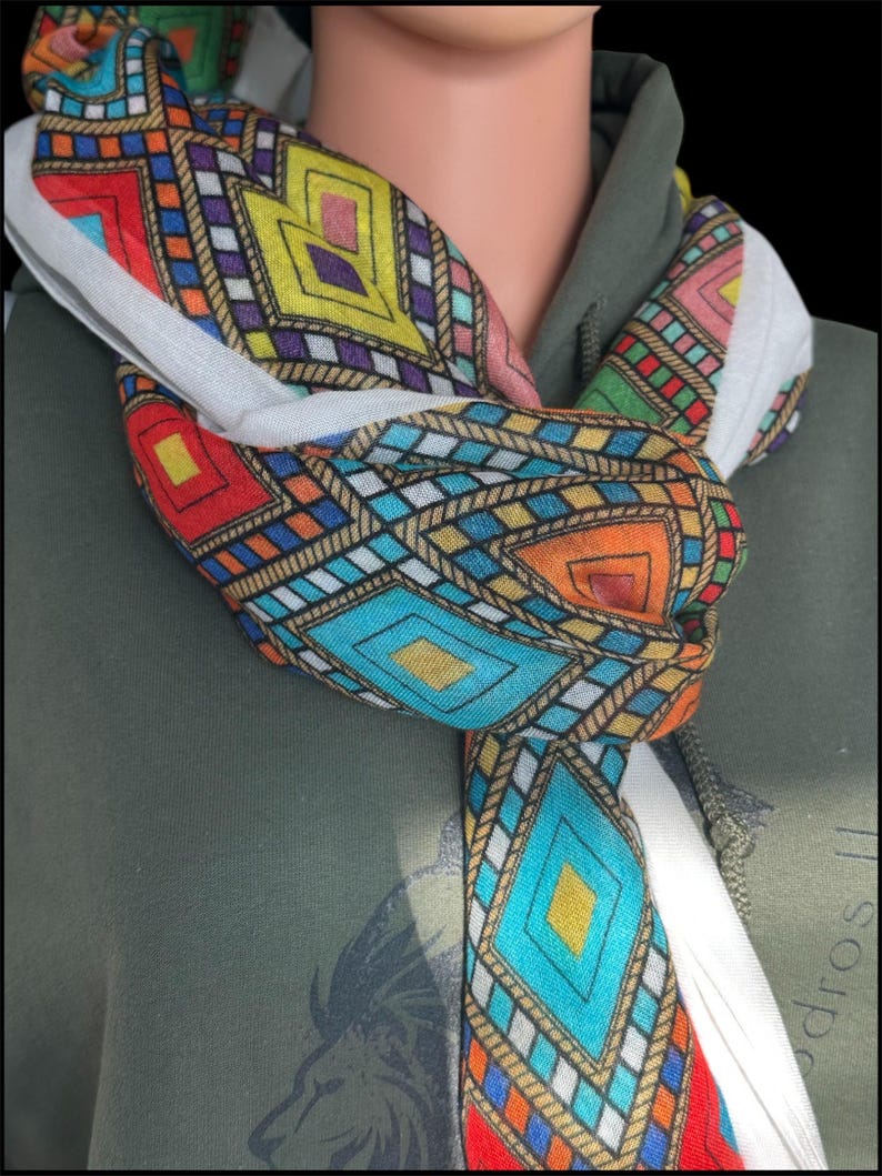 May include: A colorful scarf with a geometric pattern in red, orange, yellow, blue, and purple is knotted around a neck. The scarf has a white border and is draped over a green hooded sweatshirt. The pattern includes diamond and square shapes.