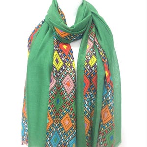May include: A green scarf with a vibrant geometric pattern. The scarf is displayed on a mannequin, highlighting its design. The pattern incorporates diamond shapes in shades of orange, yellow, and blue, adding a colourful touch to the accessory.