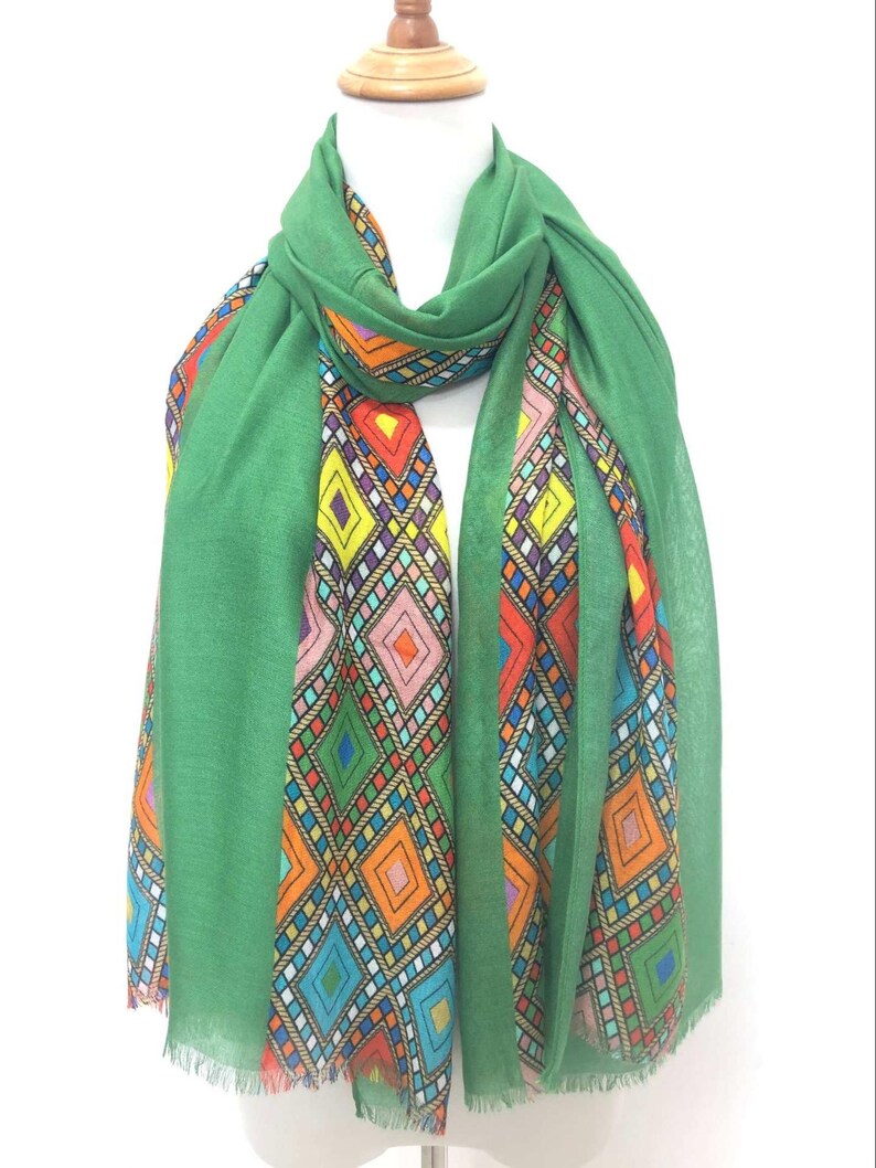 May include: A green scarf with a vibrant geometric pattern. The scarf is displayed on a mannequin, highlighting its design. The pattern incorporates diamond shapes in shades of orange, yellow, and blue, adding a colourful touch to the accessory.