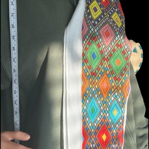 May include: A colorful, patterned scarf with a white border is draped over a green hoodie. The scarf features a geometric design with diamonds in various colors, including red, blue, and yellow. A measuring tape shows a length of approximately 34 inches.