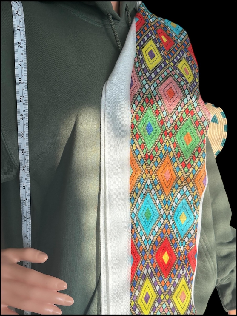 May include: A colorful, patterned scarf with a white border is draped over a green hoodie. The scarf features a geometric design with diamonds in various colors, including red, blue, and yellow. A measuring tape shows a length of approximately 34 inches.