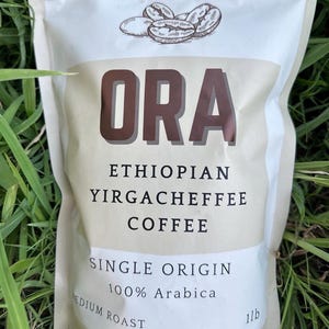 May include: A beige coffee bag with the word "ORA" in large brown letters. The bag features the text "Ethiopian Yirgacheffee Coffee", "Single Origin", "100% Arabica", and "Medium Roast". A drawing of coffee beans is at the top. The bag contains 454 grams of coffee.