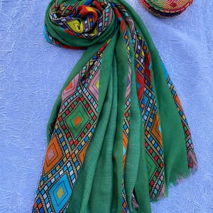May include: A green scarf with vibrant geometric patterns in orange, blue, red, and yellow. The scarf is made of a soft material and has a fringed edge. A matching woven hat is also visible.