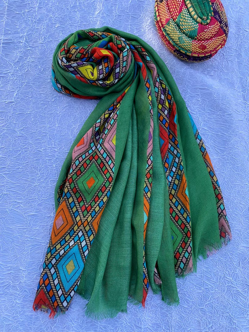 May include: A green scarf with vibrant geometric patterns in orange, blue, red, and yellow. The scarf is made of a soft material and has a fringed edge. A matching woven hat is also visible.