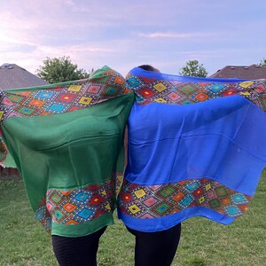 May include: Two colourful scarves, one green and one blue, are held up against a backdrop of a cloudy sky. Each scarf features a vibrant geometric pattern in shades of red, orange, yellow, and blue along the border. The scarves are made of a lightweight fabric.