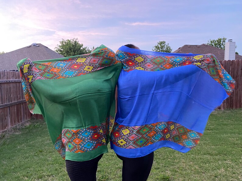 May include: Two colourful scarves, one green and one blue, are held up against a backdrop of a cloudy sky. Each scarf features a vibrant geometric pattern in shades of red, orange, yellow, and blue along the border. The scarves are made of a lightweight fabric.