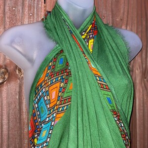 May include: A green scarf with a vibrant geometric pattern. The scarf is displayed on a mannequin, highlighting the design. The pattern features diamonds and rectangles in orange, blue, yellow, and red.