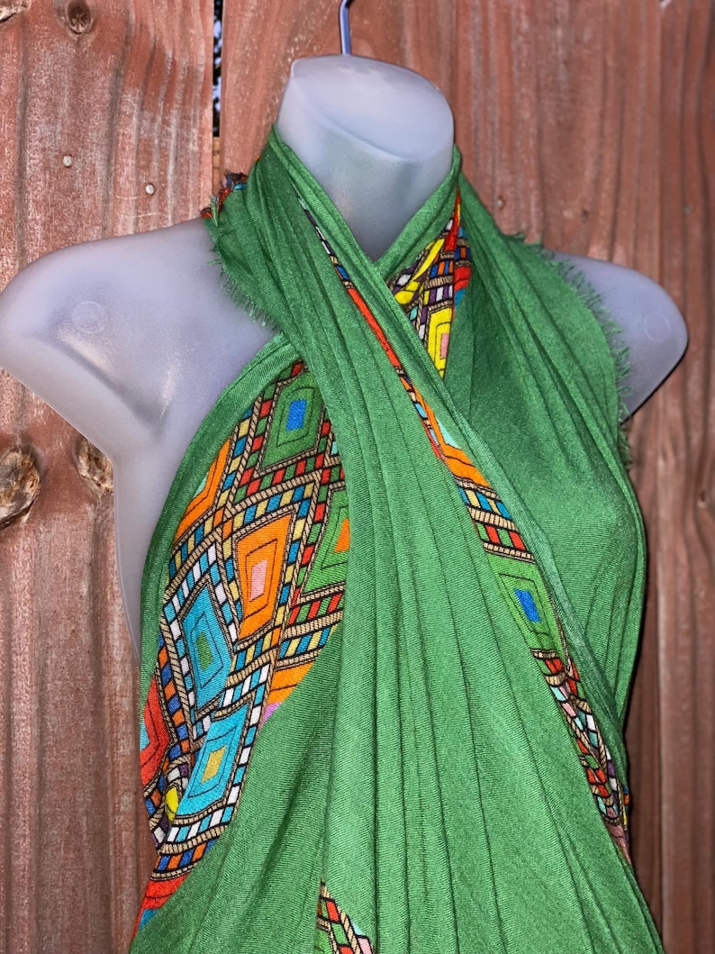 May include: A green scarf with a vibrant geometric pattern. The scarf is displayed on a mannequin, highlighting the design. The pattern features diamonds and rectangles in orange, blue, yellow, and red.