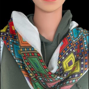 May include: A colorful infinity scarf with a geometric pattern. The scarf features a white background with a repeating design of diamonds and rectangles in shades of red, blue, green, yellow, and orange. The scarf is wrapped around a mannequin.