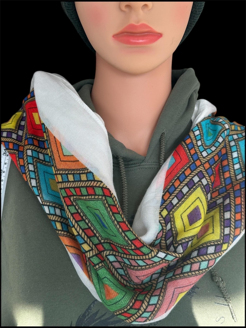 May include: A colorful infinity scarf with a geometric pattern. The scarf features a white background with a repeating design of diamonds and rectangles in shades of red, blue, green, yellow, and orange. The scarf is wrapped around a mannequin.