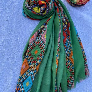 May include: A green scarf with a vibrant geometric design. The scarf features a fringed edge and is displayed on a white surface. The pattern incorporates diamond and rectangular shapes in orange, blue, pink, and yellow. A woven basket is visible in the background.