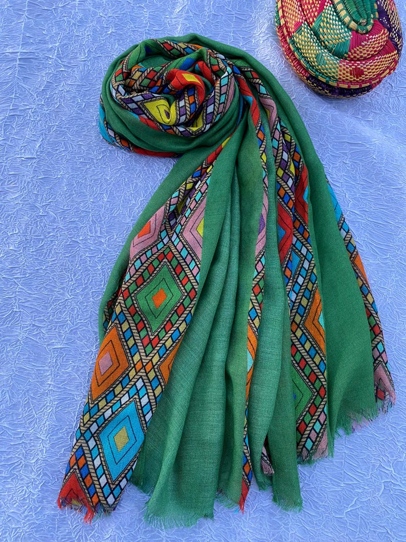 May include: A green scarf with a vibrant geometric design. The scarf features a fringed edge and is displayed on a white surface. The pattern incorporates diamond and rectangular shapes in orange, blue, pink, and yellow. A woven basket is visible in the background.