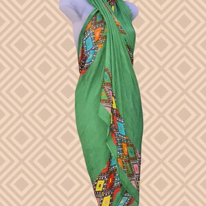 May include: A green sarong with a colourful geometric pattern. The sarong is draped over a mannequin, showcasing its design. The pattern features diamonds in shades of orange, blue, yellow, and red. The fabric appears lightweight and suitable for warm weather.