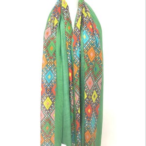 May include: A green scarf with a vibrant geometric design. The pattern incorporates diamond shapes in red, orange, yellow, blue, and pink. The scarf is displayed on a mannequin and has a fringed border.