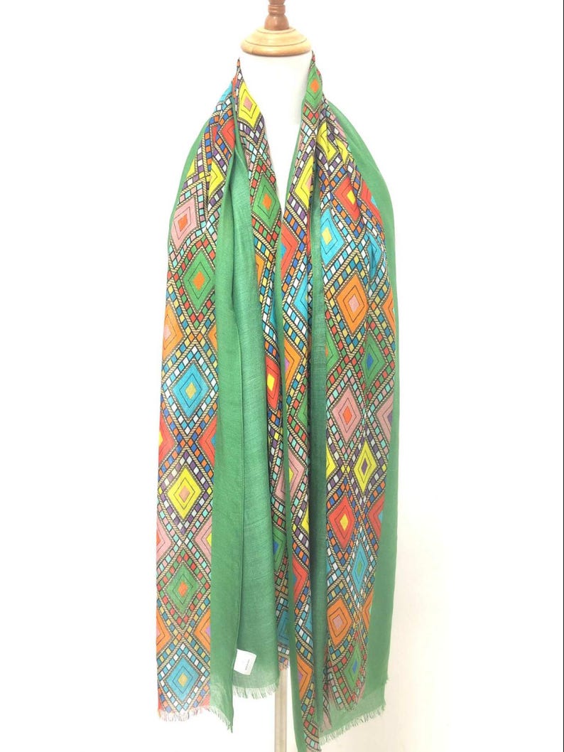 May include: A green scarf with a vibrant geometric design. The pattern incorporates diamond shapes in red, orange, yellow, blue, and pink. The scarf is displayed on a mannequin and has a fringed border.