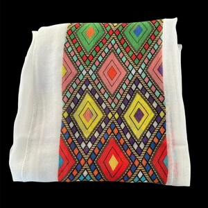 May include: White textile with a central band featuring a colorful geometric diamond pattern. The diamonds are in shades of red, yellow, green, pink, and blue, outlined with black and brown. The textile is folded and appears to be made of a lightweight fabric.